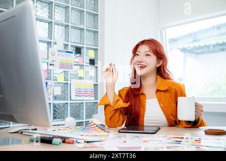 A beautiful, creative Asian female graphic designer with red curly hair is enjoying a coffee while working in her studio, sitting at a table, surround Stock Photo