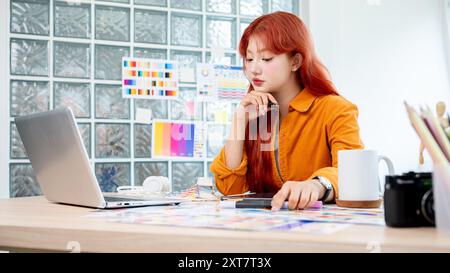 A beautiful and creative Asian female graphic designer with red curly hair is working in her creative studio, focusing on selecting colors from a set Stock Photo