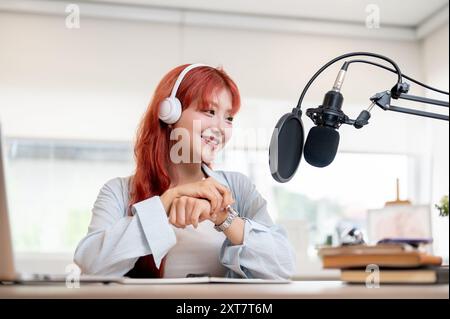 An attractive and confident Asian female podcaster or radio host with red hair is recording a new episode for her podcast channel, working in her home Stock Photo