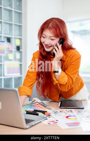 A confident Asian female graphic designer with red curly hair is talking on the phone while leaning against the table and working on her laptop, worki Stock Photo