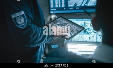 Female security officer talks with African American male coworker, uses tablet computer with surveillance footage. Diverse CCTV operators control security cameras on monitors in monitoring center. Stock Photo