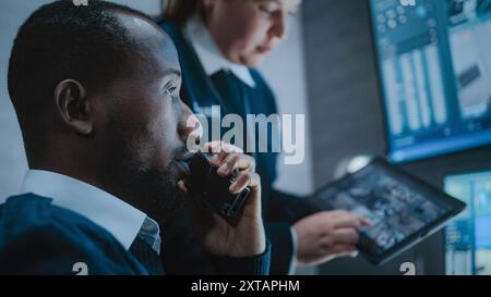 Multiethnic CCTV operators control security cameras on screens in monitoring center. Female worker talks with African American male security officer, uses tablet computer with surveillance footage. Stock Photo