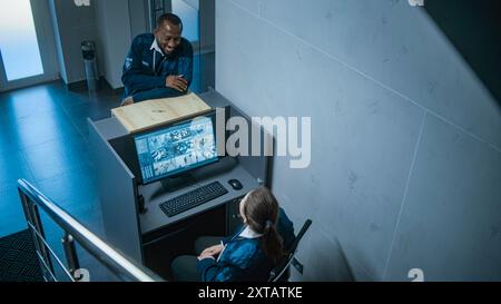 Caucasian female security guard sits at the workplace in the office working on computer with real time view from surveillance cameras. African American employee gives keys to coworker. CCTV system. Stock Photo
