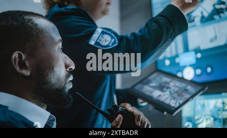Multiethnic CCTV operators control security cameras on screens in monitoring center. Female worker talks with African American male security officer, uses tablet computer with surveillance footage. Stock Photo