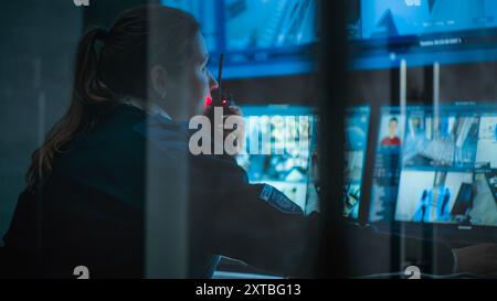 Close up of female CCTV operator working on computer in monitoring center with security cameras, talking on walkie talkie. Multiple monitors and big digital screen showing surveillance video footage. Stock Photo