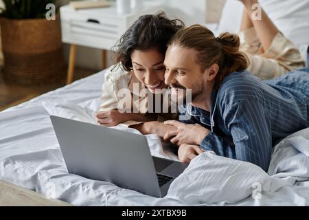 A couple is lying on a bed, looking at a laptop and smiling. Stock Photo