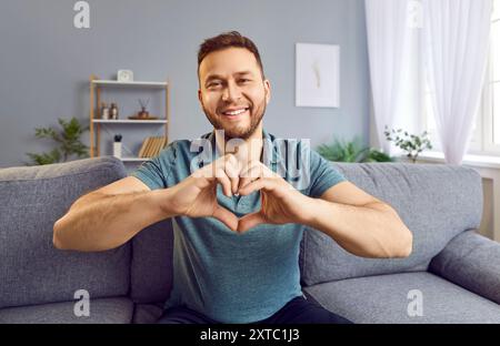 Young man sitting at home and showing love sign with hands making heart and looking at camera. Stock Photo