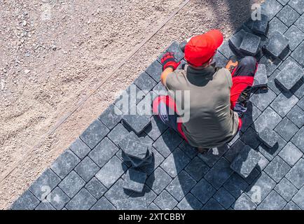 A worker in a red cap and gloves carefully places paving stones on a construction site under clear skies. Stock Photo