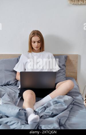 A young woman sits on a bed, using a laptop, surrounded by soft blankets. Stock Photo