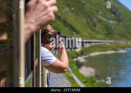 A woman leans out of the open window of the Alpine Classic Pullman Express, photographing the passing lakeside scenery at the Oberalpsee with her smartphone, while a fellow passenger holds onto the window frame in the foreground. The train dates from 1931 and originally ran as the 'Golden Mountain Pullman Express' on the Montreux–Bernese Oberland railway line. The Rhaetian Railway bought it in 1939. Uri, Switzerland. Stock Photo