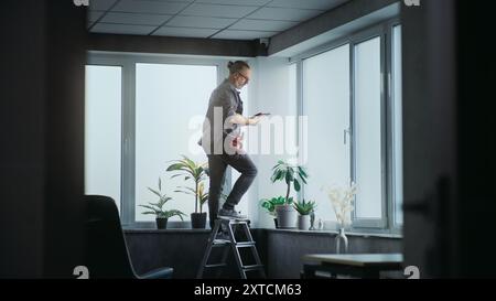 Professional male installer in uniform stands on ladder and screws surveillance camera to the ceiling in office room using tools. Monitoring and tracking. CCTV system, security and privacy concept. Stock Photo