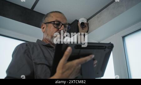 Male mature technician adjusts angle of security camera in office room using tablet computer and professional software. Monitoring and tracking system. Surveillance system, CCTV and privacy concept. Stock Photo