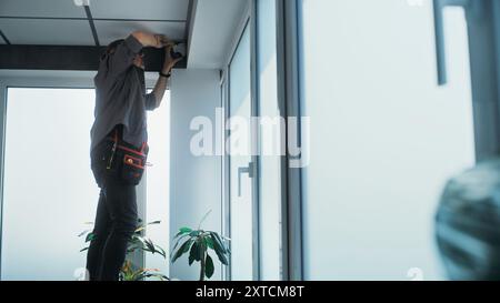 Professional male technician in uniform installs and screws security camera to the ceiling in office room using tools. Monitoring and tracking system. Surveillance system, CCTV and privacy concept. Stock Photo