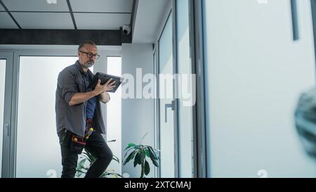 Male mature technician stands on ladder, installs CCTV camera in office room using tablet computer with professional software. Monitoring and tracking. Security system, video surveillance and privacy. Stock Photo