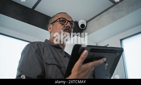Male mature technician adjusts angle of security camera in office room using tablet computer and professional software. Monitoring and tracking system. Surveillance system, CCTV and privacy concept. Stock Photo