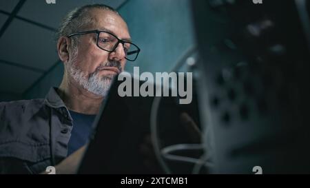 Close up of mature specialist inserting cable into server rack. Network administrator works in server room using tablet computer. Operation of information systems and networks. Data storage center. Stock Photo