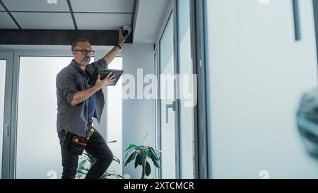 Male mature technician stands on ladder, installs CCTV camera in office room using tablet computer with professional software. Monitoring and tracking. Security system, video surveillance and privacy. Stock Photo