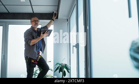 Male mature technician stands on ladder, installs CCTV camera in office room using tablet computer with professional software. Monitoring and tracking. Security system, video surveillance and privacy. Stock Photo