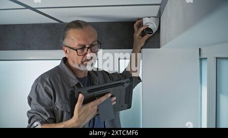 Male Caucasian technician uses tablet computer with professional software to set up security camera in office room, adjusts the angle. Monitoring and tracking system. Surveillance, CCTV and privacy. Stock Photo