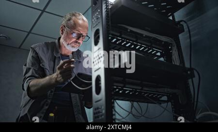 Mature engineer inserts the cable into the communications equipment, uses tablet computer. Male system administrator works in server room. Local area network or web server maintenance. Tech support. Stock Photo