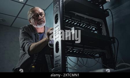 Mature engineer inserts the cable into the communications equipment, uses tablet computer. Male system administrator works in server room. Local area network or web server maintenance. Tech support. Stock Photo