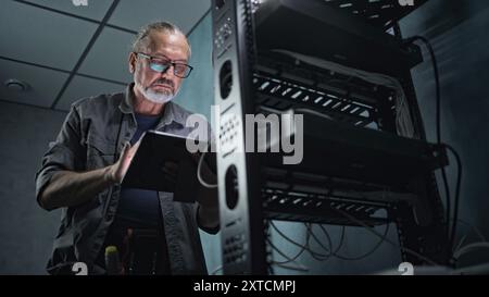 Mature engineer inserts the cable into the communications equipment, uses tablet computer. Male system administrator works in server room. Local area network or web server maintenance. Tech support. Stock Photo