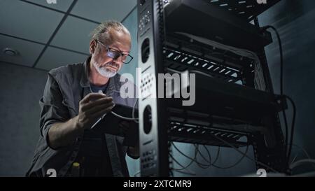 Mature engineer inserts the cable into the communications equipment, uses tablet computer. Male system administrator works in server room. Local area network or web server maintenance. Tech support. Stock Photo