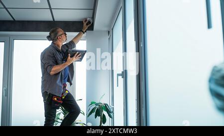 Male mature technician stands on ladder, installs CCTV camera in office room using tablet computer with professional software. Monitoring and tracking. Security system, video surveillance and privacy. Stock Photo