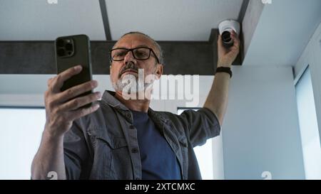 Male mature technician in uniform sets up CCTV camera in office building using mobile phone and professional app. Monitoring and tracking system. Surveillance system, security and privacy concept. Stock Photo