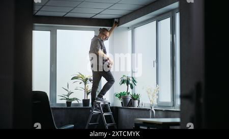 Professional male installer in uniform stands on ladder and screws surveillance camera to the ceiling in office room using tools. Monitoring and tracking. CCTV system, security and privacy concept. Stock Photo