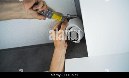 Professional male technician or installer attaches security camera to the ceiling in office building using screwdriver tool. Monitoring and tracking system. CCTV system, video surveillance and privacy. Stock Photo