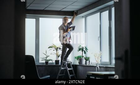 Male mature technician stands on ladder, installs CCTV camera in office room using tablet computer with professional software. Monitoring and tracking. Security system, video surveillance and privacy. Stock Photo