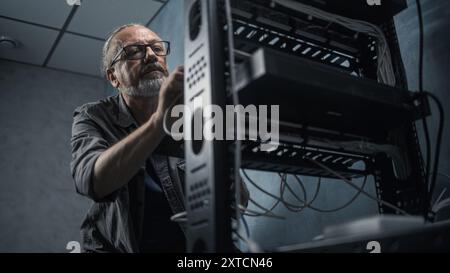 Mature engineer inserts the cable into the communications equipment, uses tablet computer. Male system administrator works in server room. Local area network or web server maintenance. Tech support. Stock Photo