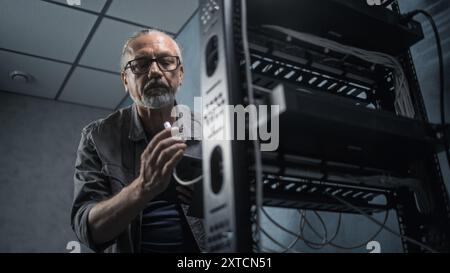 Mature engineer inserts the cable into the communications equipment, uses tablet computer. Male system administrator works in server room. Local area network or web server maintenance. Tech support. Stock Photo