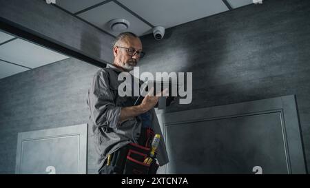 Male installer adjusts the angle of CCTV camera in office, sets up it using digital tablet computer with professional program. Monitoring and tracking. Security system, video surveillance and privacy. Stock Photo