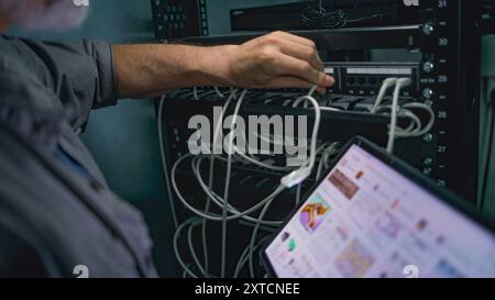Male network administrator works in server room using tablet computer. Mature specialist inserts cable into server rack. Maintenance of computer networks or web servers. Data storage center. Handheld. Stock Photo
