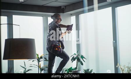 Male mature technician stands on ladder, installs CCTV camera in office room using tablet computer with professional software. Monitoring and tracking. Security system, video surveillance and privacy. Stock Photo