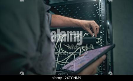 Male specialist inserts cable into server rack. Network administrator works in server room using tablet computer. Maintenance of computer networks or web servers. Data storage center. Slow motion. Stock Photo