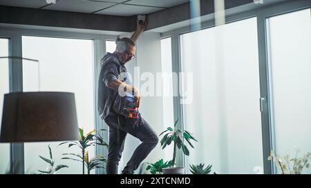 Male mature technician stands on ladder, installs CCTV camera in office room using tablet computer with professional software. Monitoring and tracking. Security system, video surveillance and privacy. Stock Photo