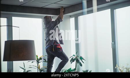 Male mature technician stands on ladder, installs CCTV camera in office room using tablet computer with professional software. Monitoring and tracking. Security system, video surveillance and privacy. Stock Photo