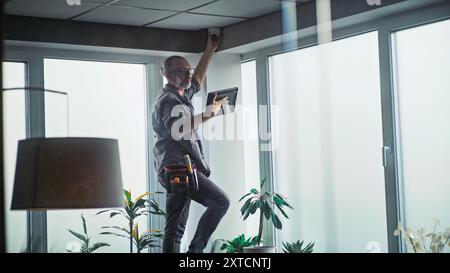 Male mature technician stands on ladder, installs CCTV camera in office room using tablet computer with professional software. Monitoring and tracking. Security system, video surveillance and privacy. Stock Photo