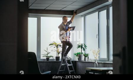 Male mature technician stands on ladder, installs CCTV camera in office room using tablet computer with professional software. Monitoring and tracking. Security system, video surveillance and privacy. Stock Photo