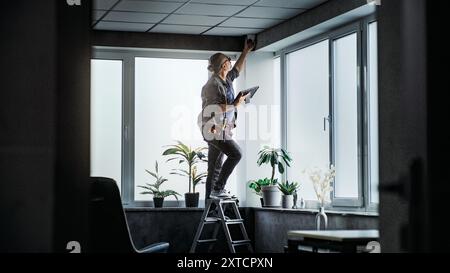 Male mature technician stands on ladder, installs CCTV camera in office room using tablet computer with professional software. Monitoring and tracking. Security system, video surveillance and privacy. Stock Photo