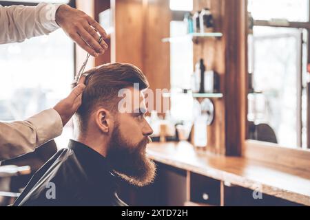 Perfect cut from back to front. Close-up side view of young bearded man getting haircut by hairdresser at barbershop Stock Photo