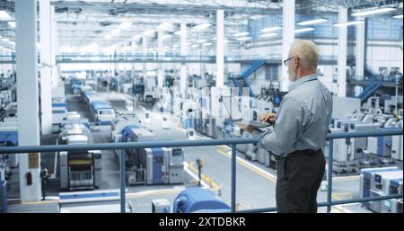 Thoughtful Middle Aged Engineer Monitoring and Analyzing Conditions at a Pristine Modern Electronics Factory with Automated Mobility Robots Working with the Help of Artificial Intelligence Software Stock Photo