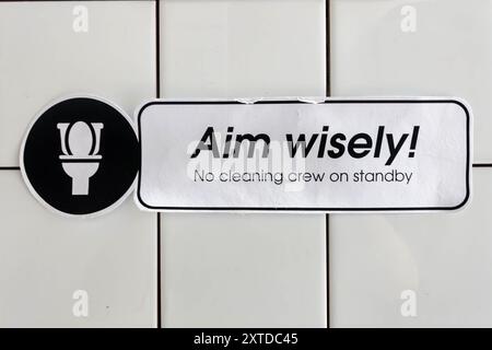 A humorous notice on a white tiled wall in a men's pubic toilets asking people to aim wisely while using the urinals. Stock Photo