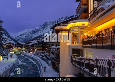 A picturesque alpine village in Zermatt is illuminated at dusk, with snow-covered mountains and a tranquil river reflecting the evening light. Stock Photo