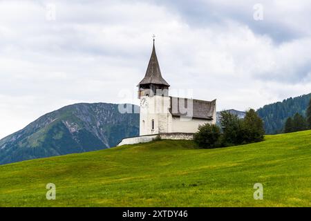 The Reformed Church of Davos Frauenkirch stands on a green knoll against the mountains of the Landwasser valley, surrounded by shrubs. Built around 1350 as the "Church of Our Dear Lady" – giving the district of Frauenkirch its name – the church was rebuilt in 1603 after a severe avalanche destroyed it on 16 January 1602, with a distinctive avalanche-splitting wedge on its uphill side that survives to this day. Landwasserstrasse, Davos, Grisons, Switzerland. Stock Photo