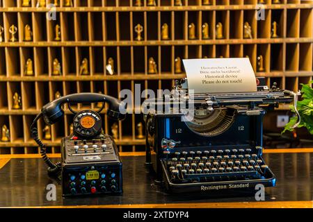 A historic Remington Standard typewriter and a rotary-dial telephone stand at the reception of the Berghotel Schatzalp, built in 1899 as a luxury sanatorium. A sign asks guests not to touch the historic devices. Behind them, numbered room-key pigeonholes fill the wall. The former Schatzalp sanatorium is now a hotel. Thomas-Mann-Weg, Davos, Grisons, Switzerland. Stock Photo