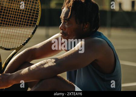African-American man with dreadlocks sitting on a tennis court holding a racket, capturing a moment of rest during a game. Stock Photo